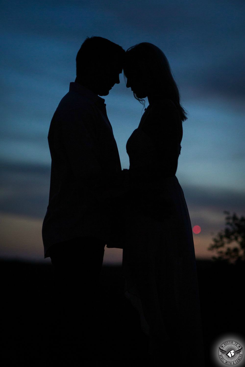 Blue and grey and orange cloudy sky in the background of a dreamy profile silhouette of a couple touching foreheads at twilight in this starry-eyed engagement photograph in Austin, Texas.
